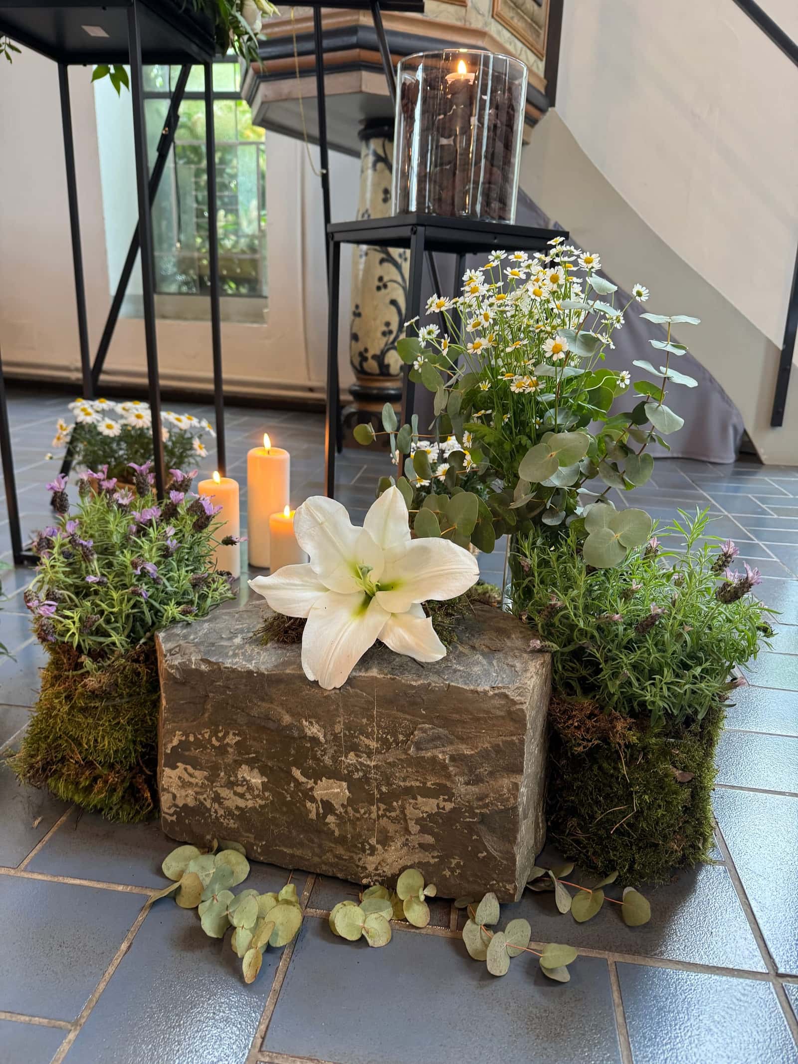 Weiße Lilie auf einem Naturstein, daneben Lavendel, Eukalyptus und brennende Kerzen