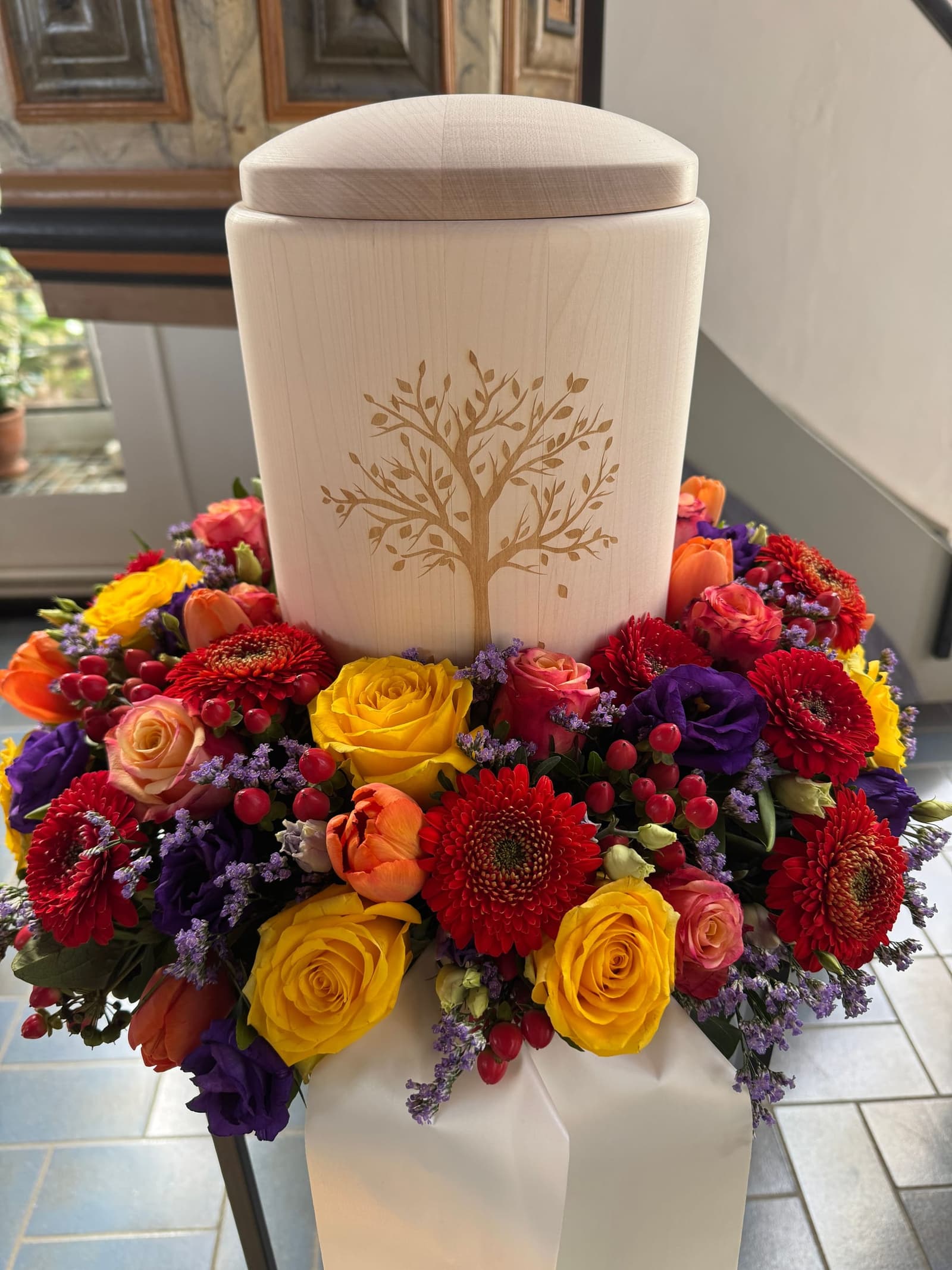 Urne aus hellem Holz mit eingraviertem Lebensbaum auf einem Kranz aus bunten Rosen und Gerbera