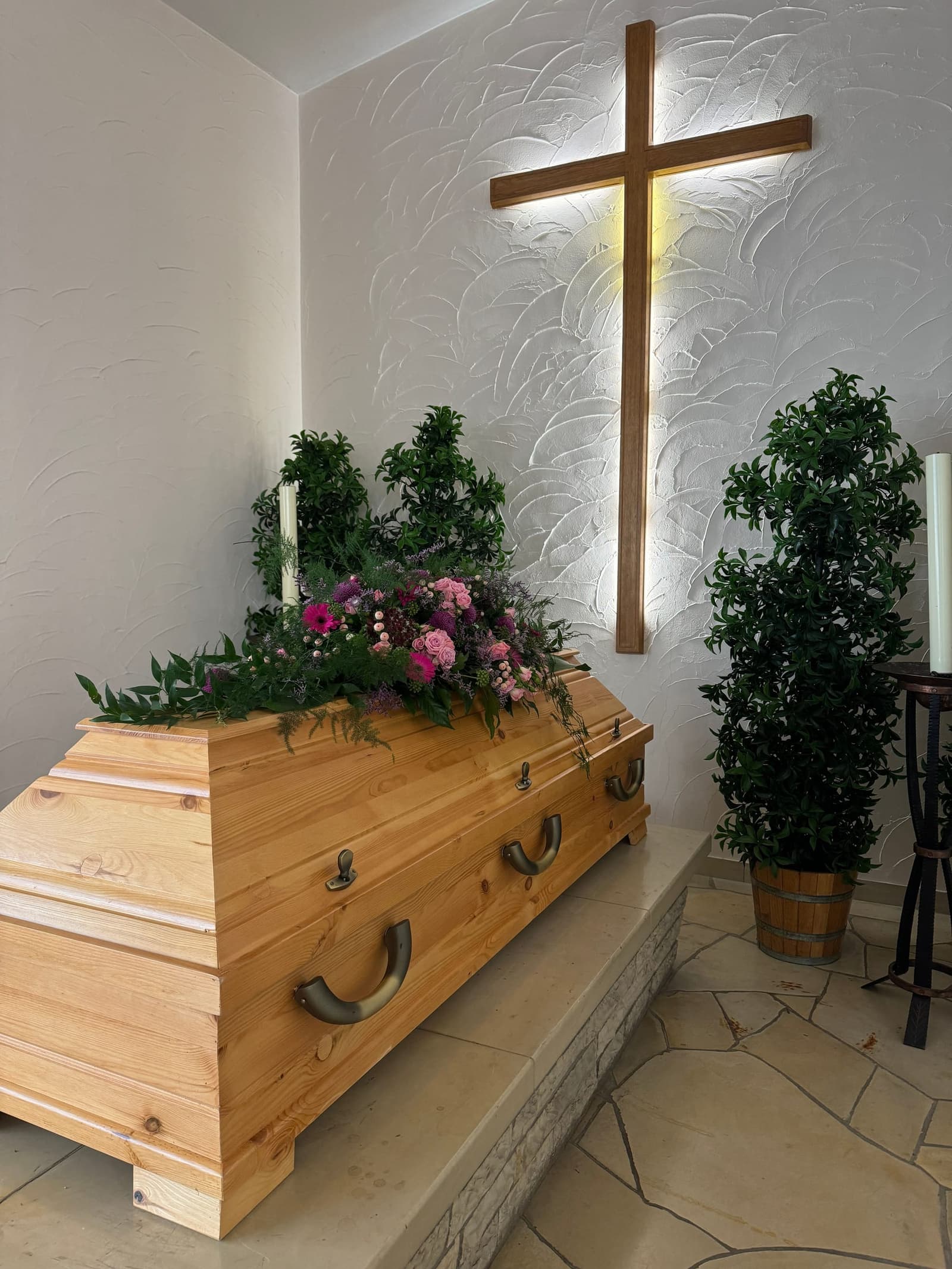 Heller Holzsarg mit einem Gesteck aus Blumen und Grün, an der Wand dahinter ein beleuchtetes Holzkreuz