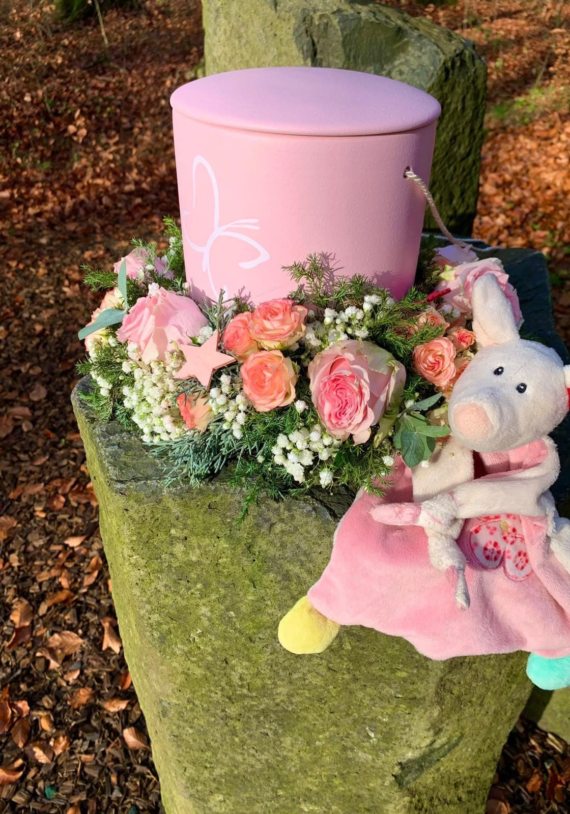 Kleine rosa Urne mit Schmetterling, umgeben von Rosen und einem Kuscheltier