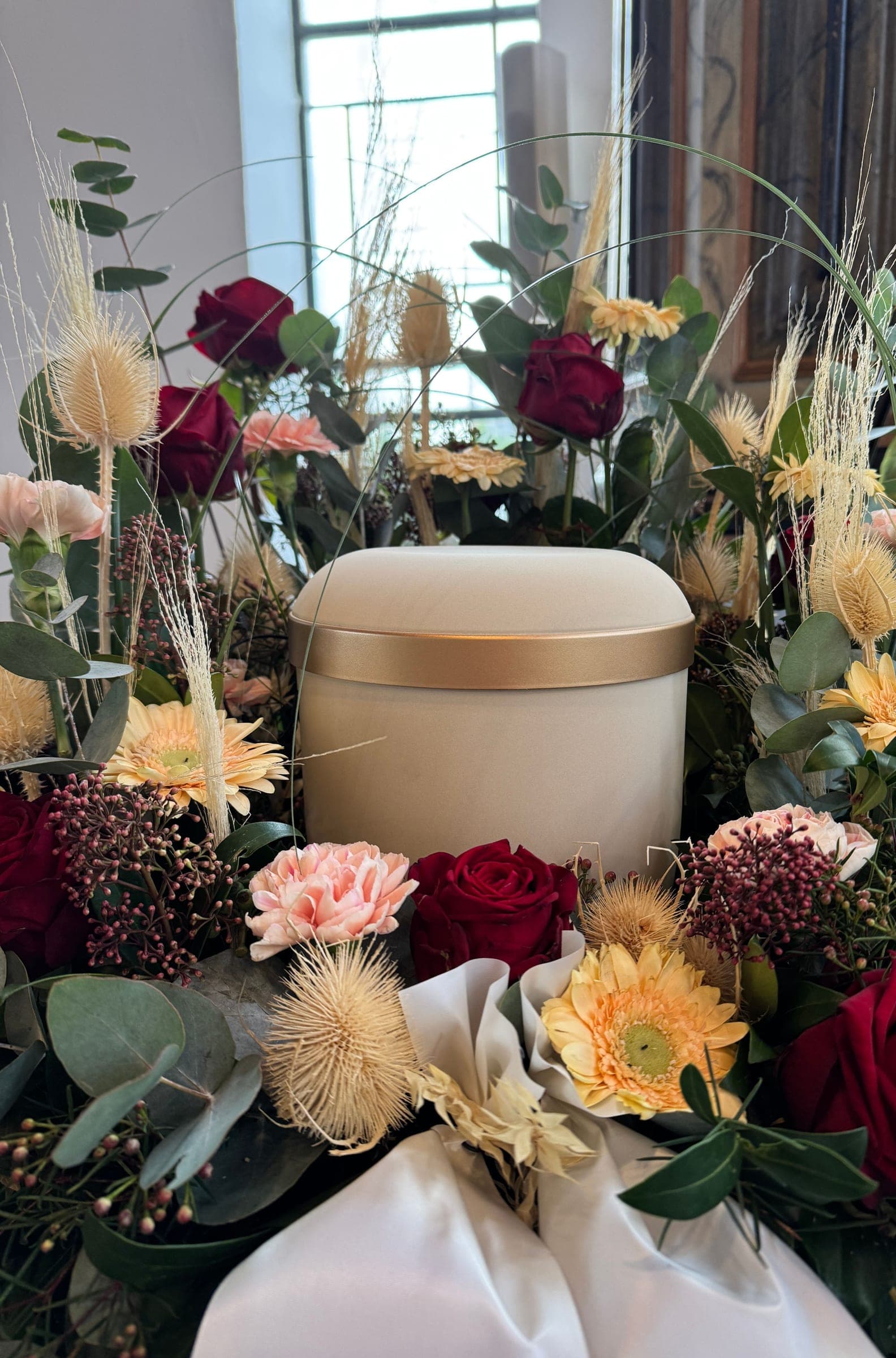 Cremefarbene Urne mit goldenem Rand zwischen roten Rosen, Gerbera und Trockenblumen