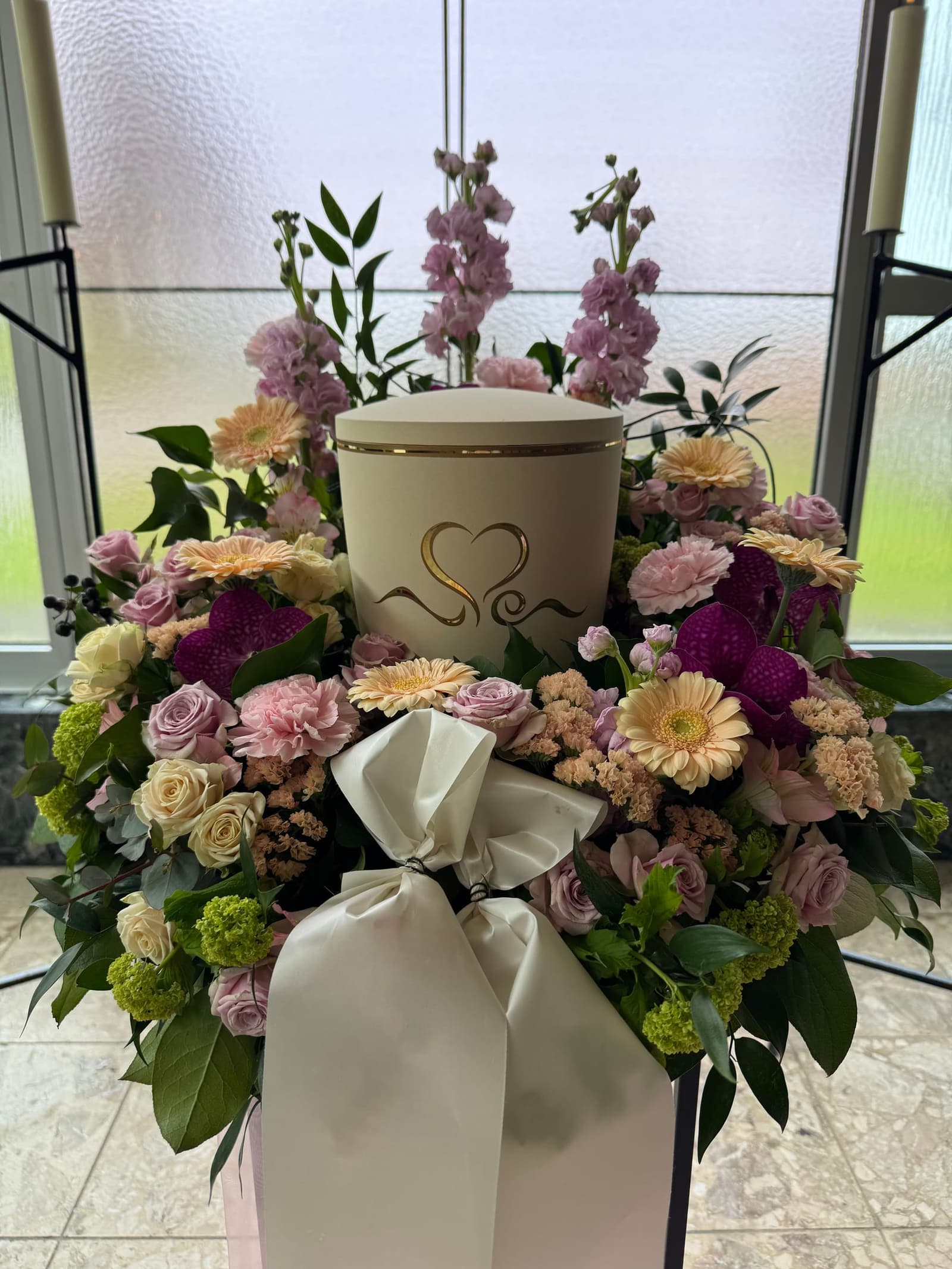 Cremefarbene Urne mit goldenem Herz, umgeben von Blumen in Rosa, Flieder und Apricot